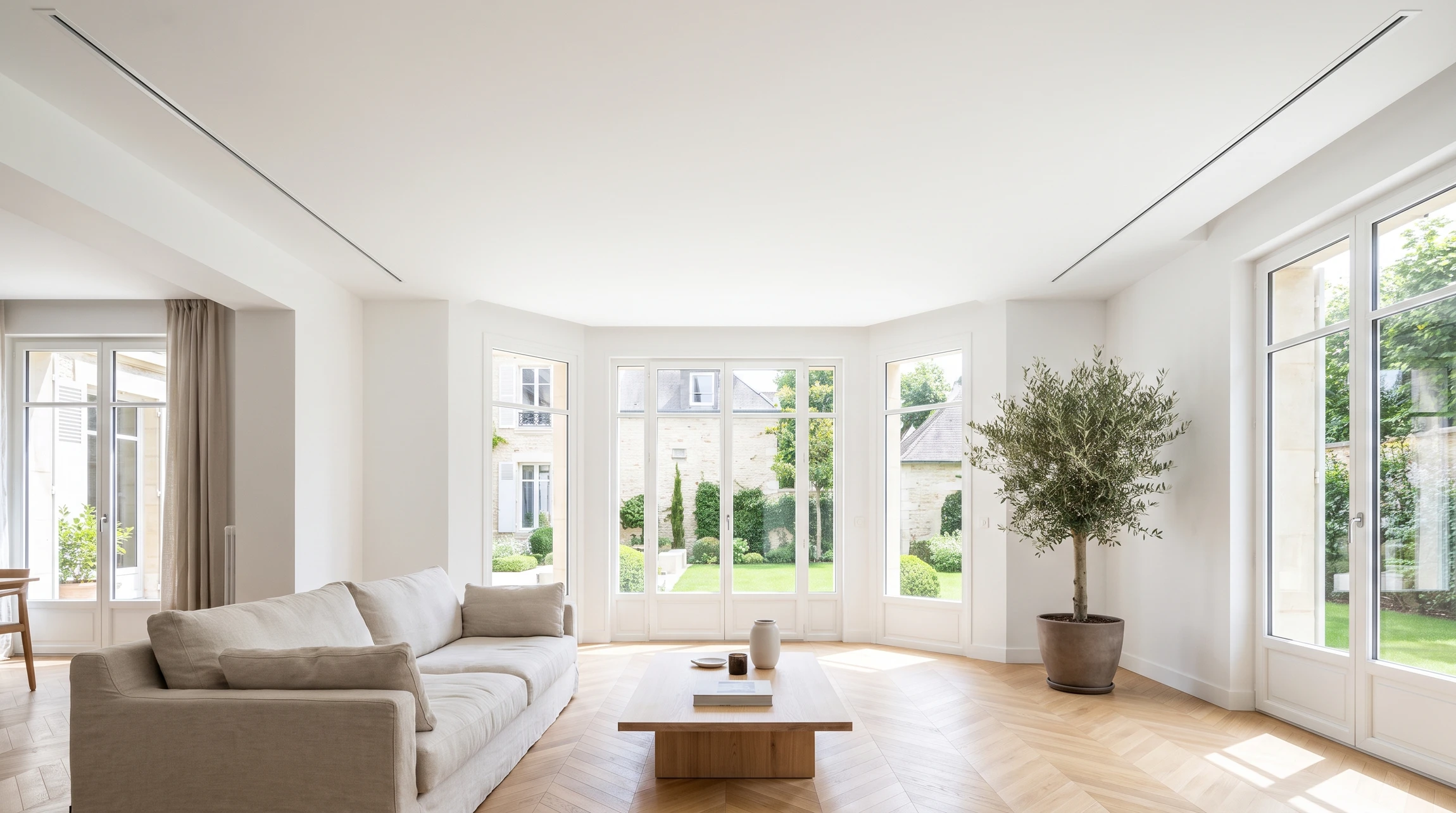 Intérieur d'une maison résidentielle moderne, pièce de vie lumineuse avec plafond blanc intégrant discrètement les gaines de climatisation gainable. Une ou plusieurs petites grilles de diffusion d'air sont visibles au plafond, pratiquement invisibles et intégrées à l'architecture. La pièce est confortable et sans appareil climatique visible mural ou au sol. Aucune conduite, compresseur ou élément technique apparent n'est visible de l'intérieur.