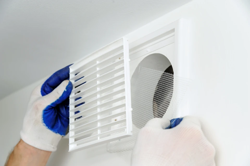 Intervention de maintenance dans un logement, technicien portant des gants de protection bleus et blancs procédant au démontage d'une grille de ventilation murale blanche