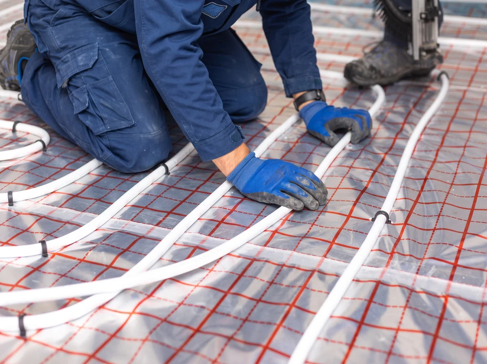 Technicien en combinaison bleue et casquette installant un radiateur mural blanc dans une pièce aux murs blancs et parquet clair